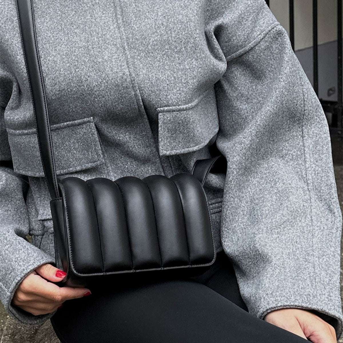 グレーのウールジャケットにブラックパンツを合わせたコーディネートに、Daniel WellingtonのSadie Bag Blackをクロスボディで取り入れたスタイリングです。縦のパフキルティングによる立体感が、シックな装いに上品なアクセントを添えます。