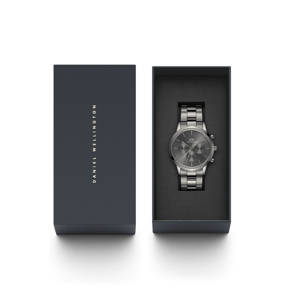 Iconic Chronograph Link Graphiteがダークネイビーのギフトボックスに収められた状態の画像です。蓋にはDaniel Wellingtonのロゴが金色で刻印されており、グラファイトカラーのクロノグラフ本体がブラックのクッション上に丁寧にセットされています。贈り物にふさわしい上質なパッケージです。
