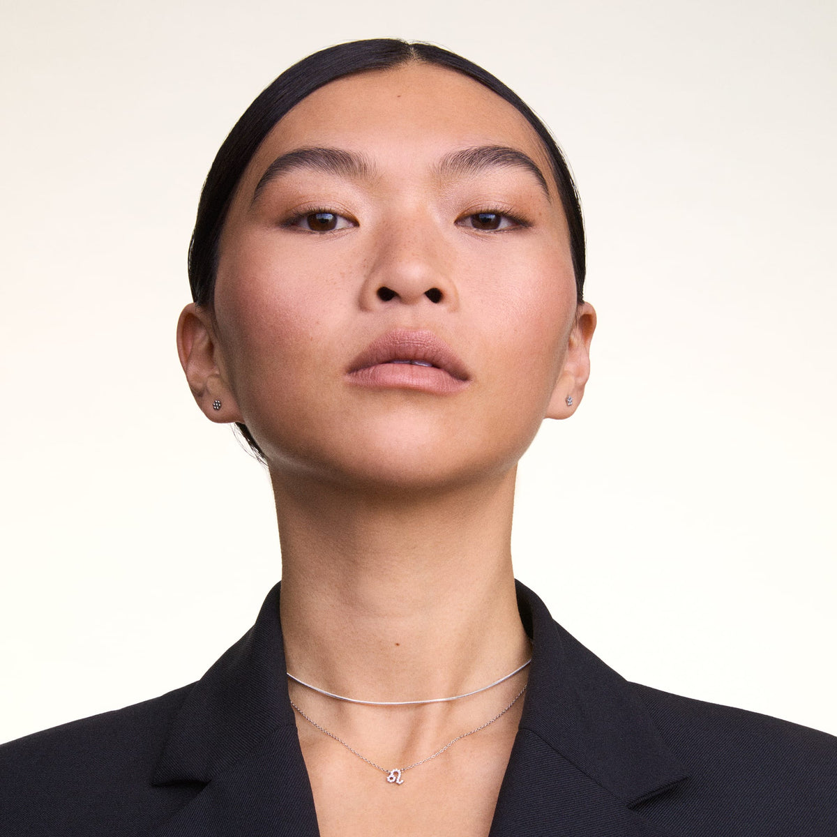 ダークネイビーのジャケットを着た女性が、Charms Snake Necklaceをチョーカースタイルで身に着け、シルバーのチャームネックレスと重ね付けした着用イメージです。