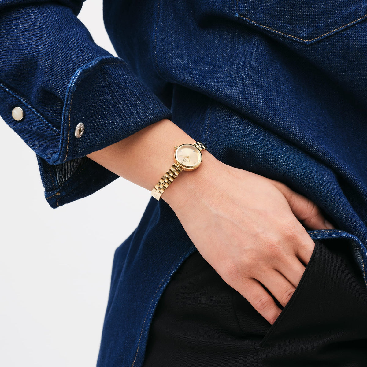Daniel Wellington Elan Lumine Unitone Goldを着用した手元のクローズアップ。デニムジャケットとブラックパンツのカジュアルなコーディネートに、ゴールドの3リンクストラップがブレスレットのように手首に自然になじんでいます。