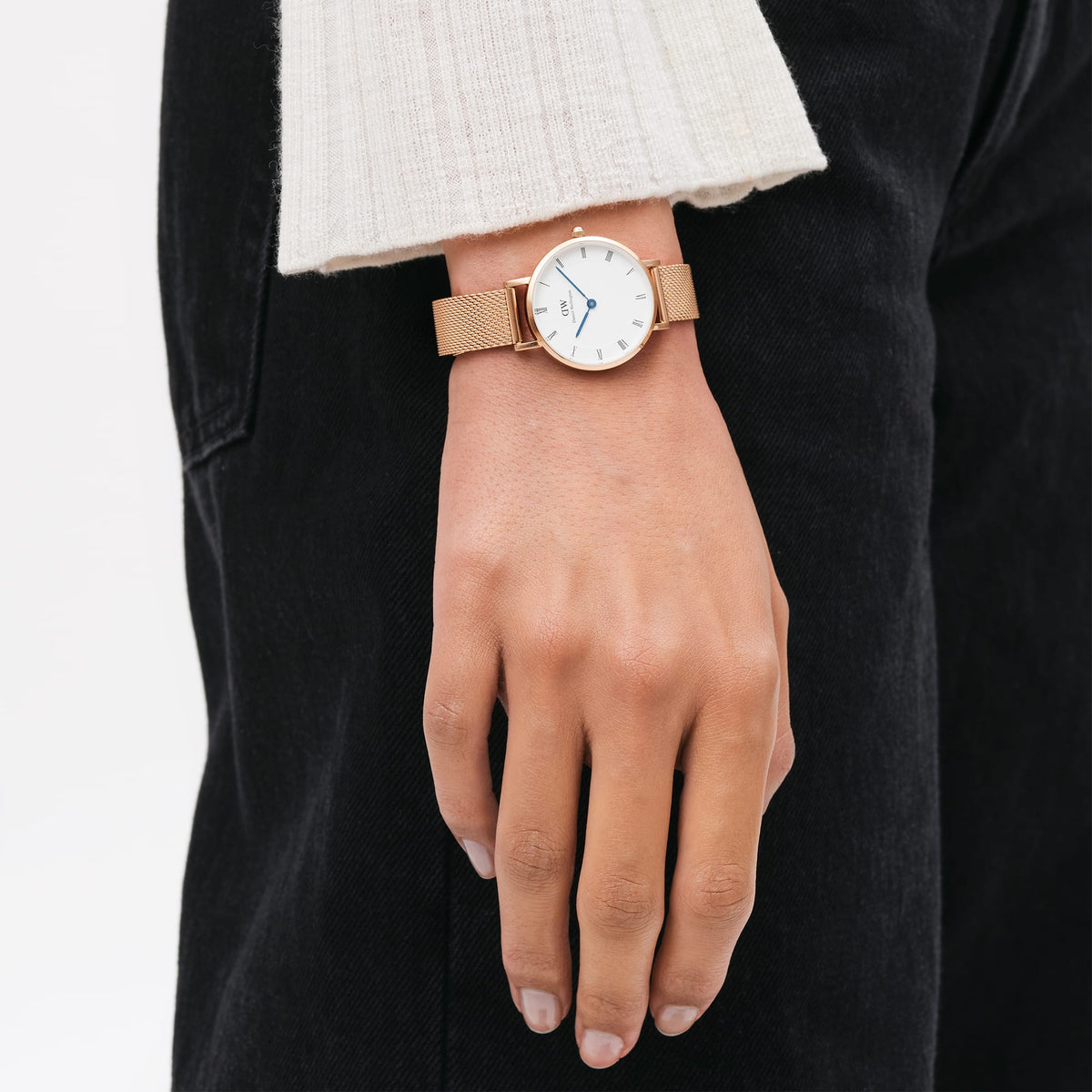 Petite Roman Numerals Melroseを手首に着用したスタイルの画像です。ベージュのニットとブラックデニムに合わせ、ローズゴールドのメッシュストラップとブルースチールの針が手元に控えめなアクセントを添えています。