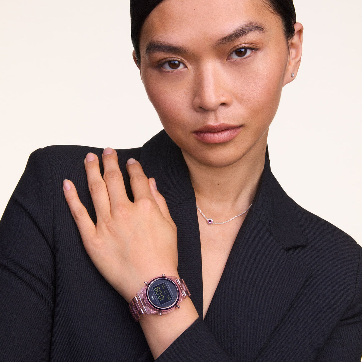 ブラックのジャケットを着用した女性が、鎖骨元でパープルのクリスタルが輝くDaniel WellingtonのFebruary Charmネックレスを身につけた着用イメージです。手首にはピンクの透明ストラップのデジタルウォッチを合わせ、スタイリッシュなコーディネートを演出しています。