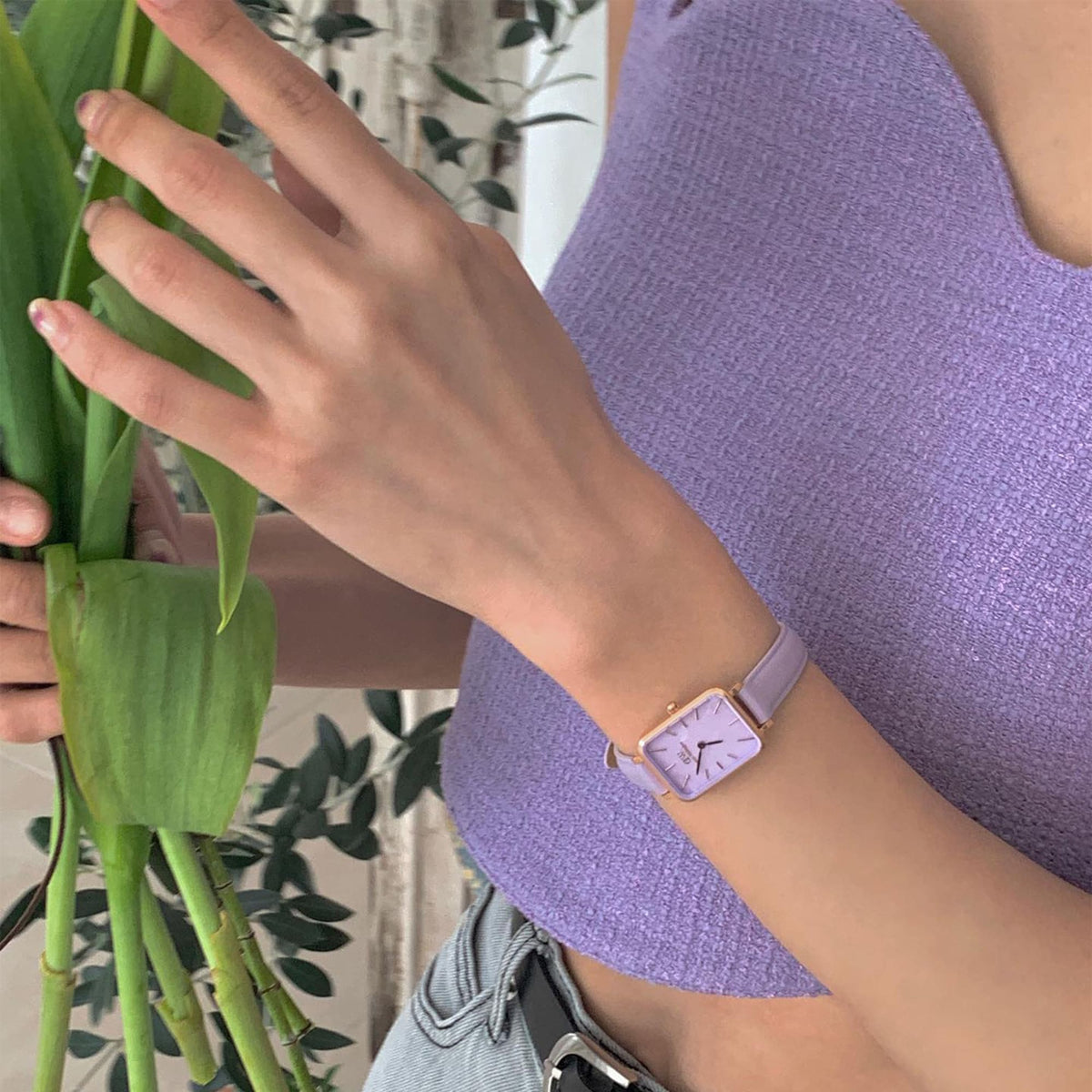 ラベンダーカラーのトップスにコーディネートした、Quadro Lavender MOP Rose Goldを着用した女性の手元です。グリーンの植物を手にしたナチュラルなスタイリングで、コンパクトなスクエアケースが手首になじんでいます。