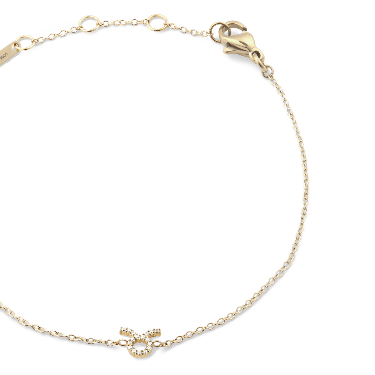 牡牛座モデルのCrystal Zodiac Chain Bracelet Goldを斜め上から撮影したクローズアップ画像です。ゴールドの繊細なチェーンが優雅な弧を描き、中央下部にはクリスタルをあしらった牡牛座のシンボルが輝いています。右上にはロブスタークラスプ、左上にはブランド刻印プレートが確認できます。