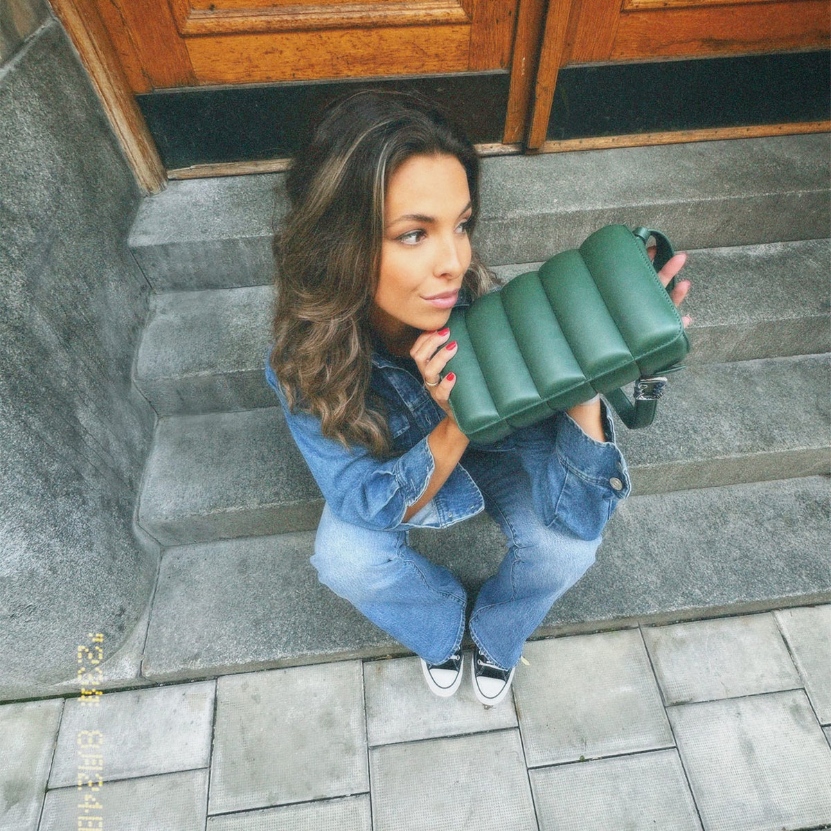デニムコーディネートの女性が石段に座り、Sadie Bag Green Sを手にしたスタイリングイメージです。縦パフ加工のグリーンレザーがカジュアルな装いに上品なアクセントを添えています。