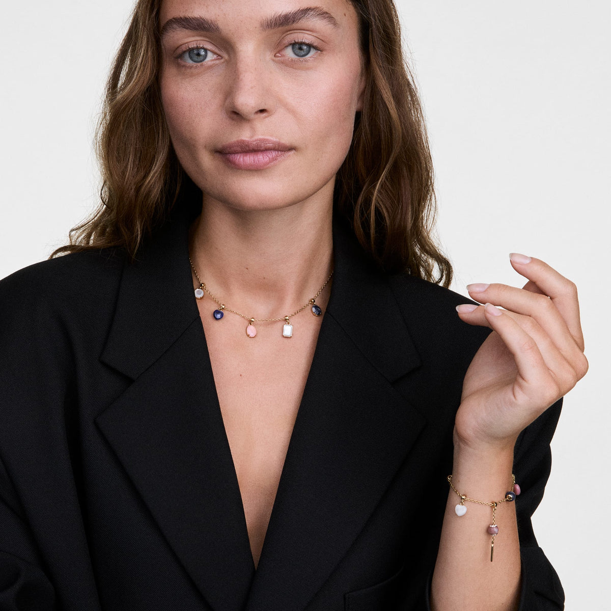 黒のジャケットを合わせた女性が、Elementalコレクションのゴールドチャームチェーンネックレスとブレスレットを着用したスタイリング例です。真珠母貝やクリスタルなど、複数のチャームを組み合わせることで、胸元と手元に華やかなアクセントを添えています。