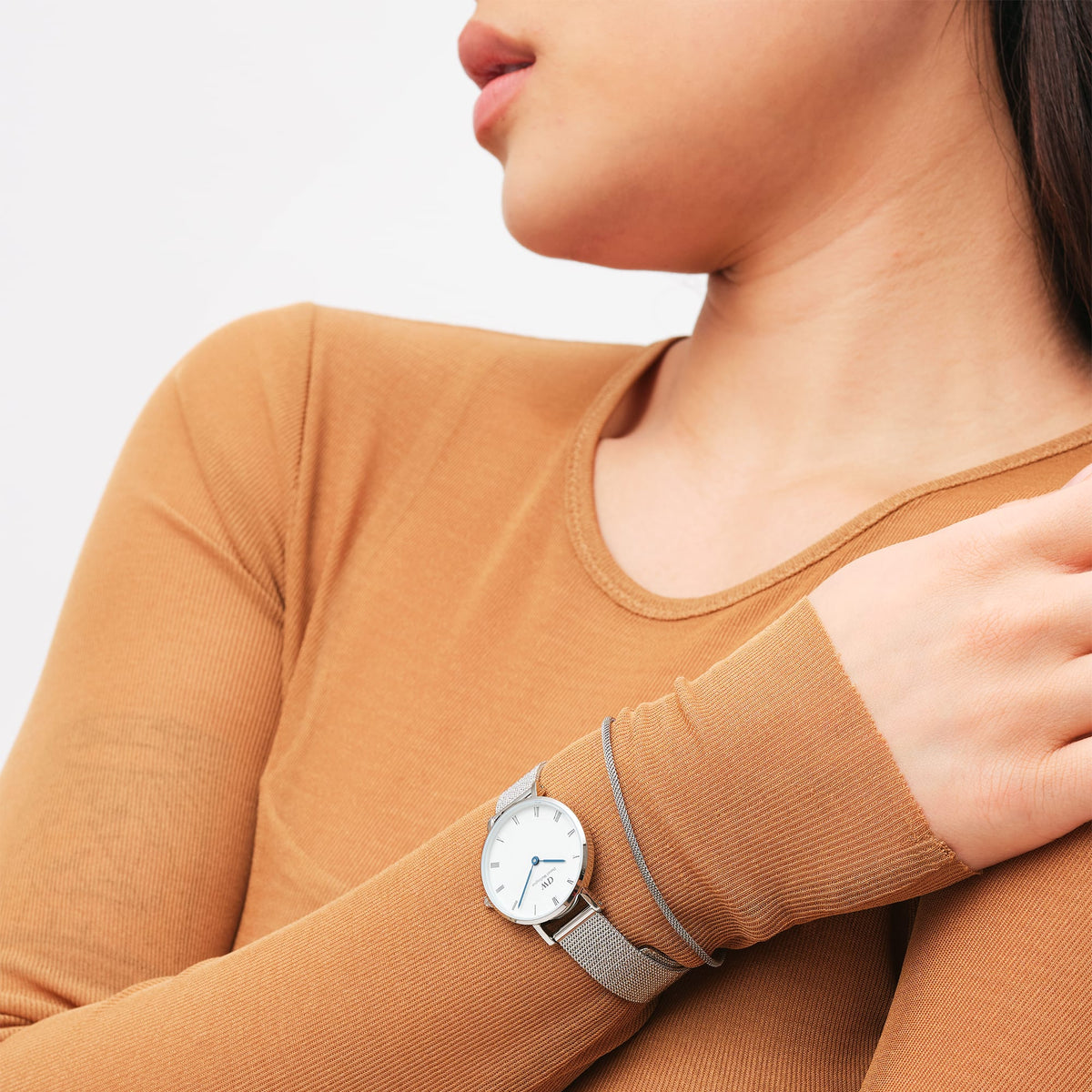 ペティットローマンニューメラルスターリングを着用した女性の手元。キャメルカラーのトップスに、シルバーメッシュストラップのコンパクトな28mmサイズの腕時計が自然になじんでいます。ブルースチールの針とローマ数字が、手首にさりげないアクセントを添えます。