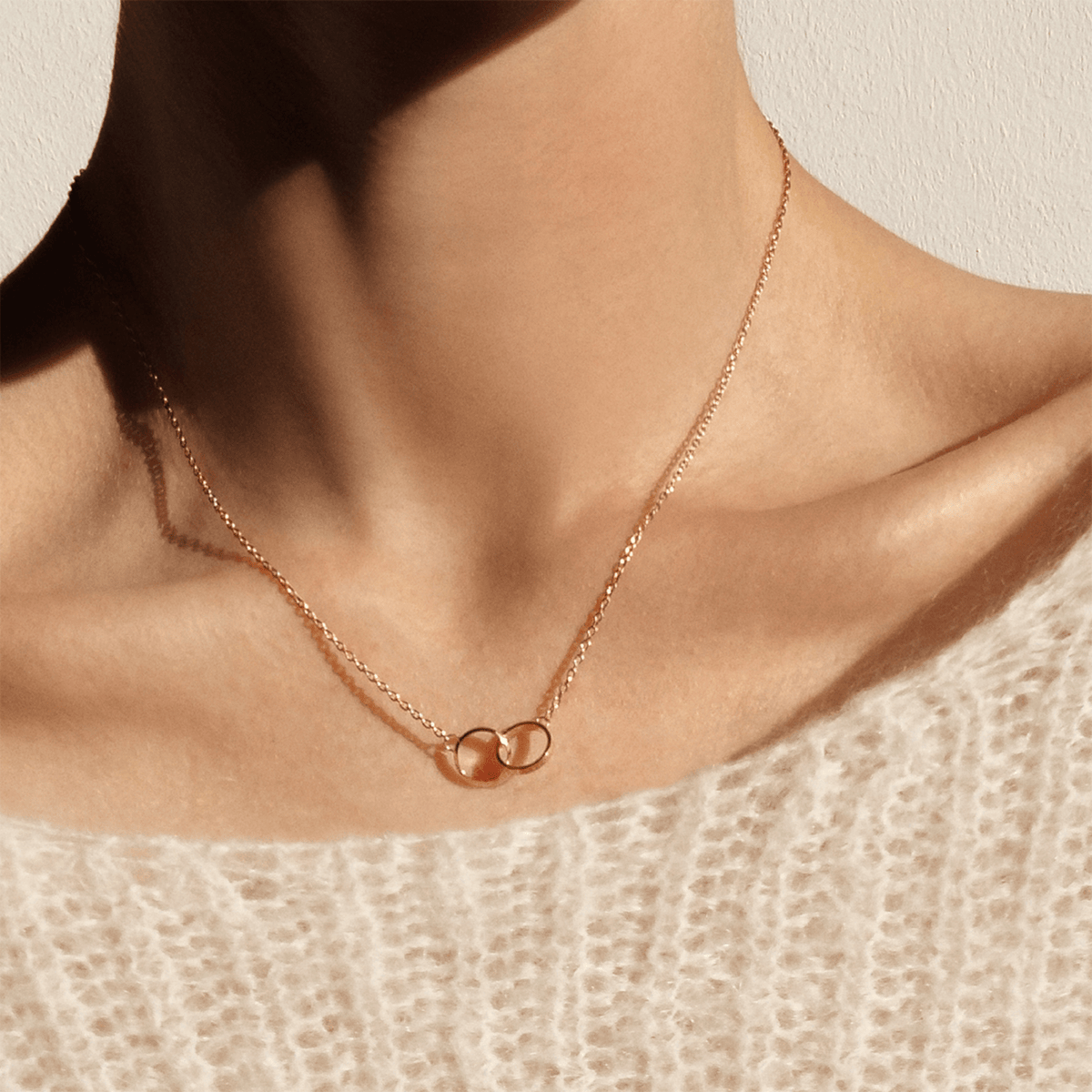 ダニエルウェリントン Elan Unity ネックレスを着用した女性の胸元。ローズゴールドのケーブルチェーンが鎖骨に沿って優雅に輝き、2つのインターロッキングリングがクリーム色のニットの上で上品に映えます。