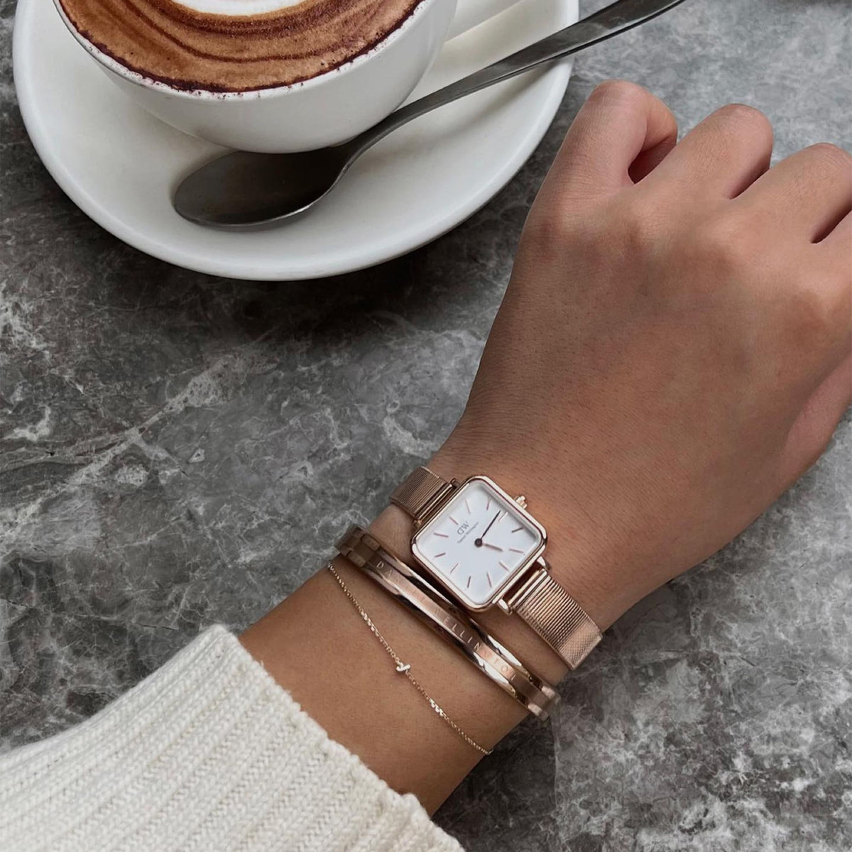 カフェでのひとときに、Daniel WellingtonのElan Braceletローズゴールドをスクエア型の腕時計やチェーンブレスレットと重ね付けした、上品な手元のスタイリング。大理石調のテーブルにカプチーノを添えた、落ち着いた空間です。
