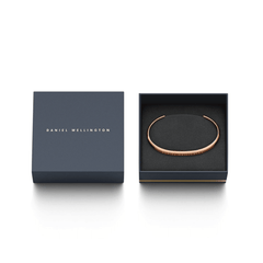 ダニエルウェリントン Classic Valentine Bracelet Rose Goldが、ネイビーの専用ギフトボックスに収められています。蓋にはゴールドのブランドロゴがあしらわれ、ローズゴールドのカフブレスレットが黒いクッショントレーの上に丁寧にセットされており、贈り物にぴったりの上品な仕上がりです。
