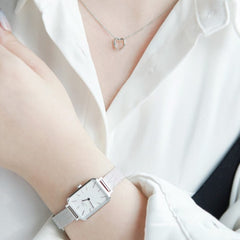 白いシャツを着た女性がElan Le Coeur Necklaceのシルバーを身に着けた様子です。小ぶりなオープンハートのペンダントが胸元で控えめに揺れ、手首にはDaniel Wellingtonのシルバーメッシュストラップの腕時計を合わせています。