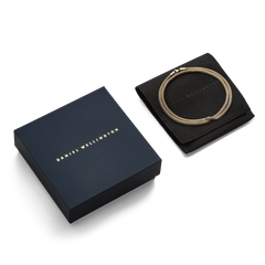 Tide Mesh Necklace Gold 2.8のギフトパッケージです。ダークネイビーのDaniel Wellingtonロゴ入りボックスの横に、ブランドロゴが型押しされたブラックのスエード調ポーチが置かれ、その上にゴールドメッシュチェーンが丸く収められています。