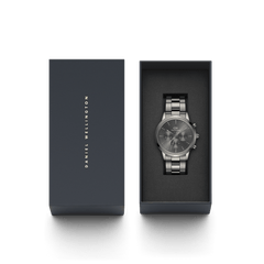 Iconic Chronograph Link Graphiteがダークネイビーのギフトボックスに収められた状態の画像です。蓋にはDaniel Wellingtonのロゴが金色で刻印されており、グラファイトカラーのクロノグラフ本体がブラックのクッション上に丁寧にセットされています。贈り物にふさわしい上質なパッケージです。
