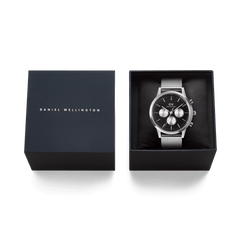 ブラックのクッショニングの上に、シルバーメッシュストラップのクロノグラフウォッチ「Iconic Chronograph Mesh Black」が丁寧にセットされたギフトボックスの画像です。ダークネイビーのボックスにはブランドロゴがあしらわれ、贈り物にもぴったりの上質な仕上がりとなっています。