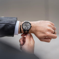 Classic Bracelet Graphiteをグラファイトカラーの腕時計と重ね付けした男性の手元。レザージャケットと白シャツの袖口から覗く細身のカフブレスレットが、さりげないアクセントとなって腕元を引き締めています。