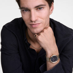 ブラックシャツを着用した男性が、Daniel WellingtonのTide Mesh Necklace Rose Gold 2.8を首元に合わせたスタイリングイメージです。ローズゴールドのメッシュウォッチやリングとコーディネートし、統一感のある上品な装いを演出しています。