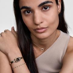 Daniel Wellington Honey Gold Setを身に着けた女性のクローズアップ。Quadro Miniのゴールドメッシュ腕時計とクリスタルテニスブレスレットを重ね付けし、胸元にはタイガーアイのAudrey Necklace、耳元にはタイガーアイのスタッドピアスを合わせた、統一感のあるコーディネートです。