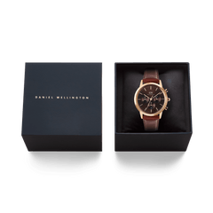 Daniel Wellington Iconic Chronograph St Mawes。ネイビーのギフトボックスに収められ、箔押しのブランドロゴが施された蓋と、クッション上にセットされたローズゴールドケース×ブラウンレザーストラップの本体が並びます。贈り物にぴったりの上品なパッケージです。
