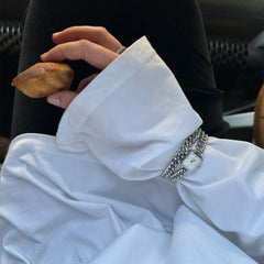 白いブラウスの袖口からのぞくJolieシルバーチェーンウォッチとChunky Chain Braceletの重ね付けスタイル。マドレーヌを手にしたカフェのシーンで、シルバーのチェーンリンクが自然光を受け、上品に輝いています。
