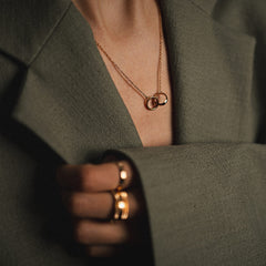 オリーブグリーンのジャケットを羽織った女性がElan Unity Necklace ローズゴールドを着けたスタイリングシーンです。胸元のVラインに沿ってインターロッキングリングが自然に収まり、指元にはローズゴールドのリングを重ねてトーンを統一しています。