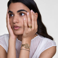 Classic Mini Signet Ring Goldを薬指に着けた女性のスタイリングイメージです。ゴールドのメッシュストラップ腕時計やブレスレットと合わせ、統一感のあるゴールドトーンのコーディネートを楽しんでいます。