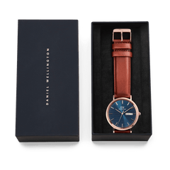 Classic Day Display St Mawes Rose Gold Arcticをネイビーのギフトボックスに収めた様子です。ブルーの文字盤とブラウンのレザーストラップが黒いクッションの上に丁寧に配置され、贈り物にぴったりの上品なパッケージングとなっています。