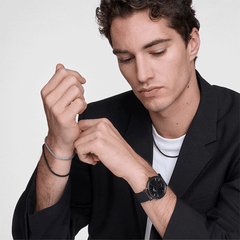 黒のジャケットを着用した男性が、メッシュブレスレットとブラックのメッシュストラップ腕時計を重ね付けしたスタイルの着用イメージです。