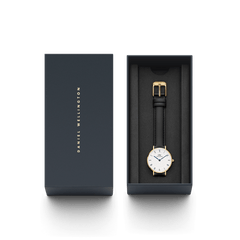 Petite Roman Numerals Sheffield Goldが、ネイビーのブランド専用ギフトボックスに収められています。ゴールドのバックルが付いたブラックレザーストラップとローマ数字の文字盤が、黒いクッションカバーの上で美しく映える、ギフトにぴったりのパッケージです。