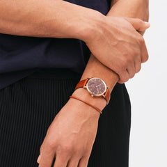 ローズゴールドのSt Mawes Braceletを、ブラウンレザーストラップのローズゴールド腕時計と重ね付けした男性の手元です。レザーブレスレットとウォッチストラップの色調が統一され、自然な一体感のあるスタイリングを演出します。