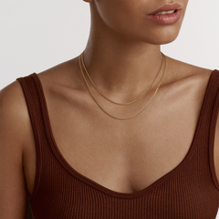 Daniel WellingtonのBox Chain Necklace Gを2本重ね付けした着用イメージです。ブラウンのタンクトップに合わせ、異なる長さのゴールドチェーンが鎖骨まわりにレイヤードスタイルを演出しています。