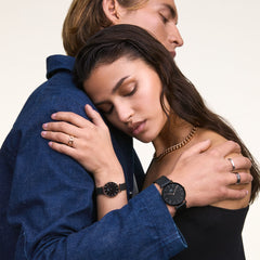 男女がブラックメッシュストラップのDaniel Wellington腕時計をペアで着用しているライフスタイル写真です。女性の手首にはPetite Ashfield Rose Goldが、ローズゴールドのリングやチェーンネックレスと合わせて上品に映えています。