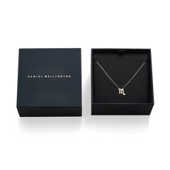 Daniel Wellingtonのネイビーギフトボックスに収められた、蠍座モチーフのCrystal Zodiac Chain Necklace Goldの俯瞰写真です。ブラックのクッション中央には、クリスタルで描かれた蠍座のシンボルをあしらったゴールドペンダントが丁寧に配置され、左側にはブランドロゴ入りの蓋が添えられています。
