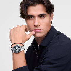 ネイビーのジャケットを着用した男性が、Daniel Wellingtonのシルバーメタルブレスレット付きクロノグラフ腕時計とブラックの編み込みレザーブレスレットを重ね付けしたスタイルの着用イメージです。