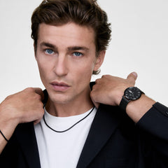 ダークジャケットと白TシャツにTide Mesh Necklace Black 1.8を合わせた、男性モデルのオールブラックコーディネート。手首にはDaniel Wellingtonのブラック腕時計を着用しています。