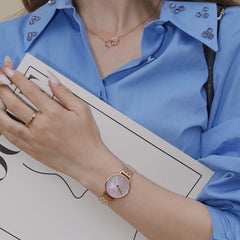 ブルーのシャツに合わせたPetite Lumine 5 Link Melroseの着用イメージです。ローズゴールドの5リンクブレスレットとピンクマザーオブパールの文字盤が、手元に上品な輝きを添えます。リングやネックレスとのコーディネートも楽しめるスタイリングです。