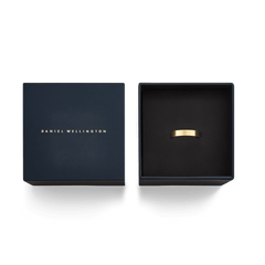 ダニエルウェリントンのLine Ring Brushed Goldが、ネイビーの専用ギフトボックスに収められた様子を真上から撮影した画像です。ゴールドのロゴがあしらわれた蓋と、黒いクッショントレーに置かれたマットゴールドのリングが、ギフトにふさわしい上品な雰囲気を演出しています。