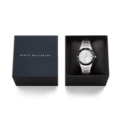 ダニエルウェリントンのIconic Paradigma Link Black Bezelが、ネイビーのブランドギフトボックスに収められた状態の画像です。箱の蓋にはダニエルウェリントンのロゴが箔押しされ、ブラックのクッション上にシルバーケースとリンクブレスレットの腕時計が丁寧にセットされています。贈り物にもぴったりの上質なパッケージです。