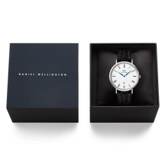 Daniel WellingtonのClassic Roman Numerals Date Black Croc Belly Silverは、ネイビーのブランドボックスに収められています。クッションの上にセットされたホワイト文字盤とブルーの針が、ギフトにも最適な上品なパッケージの中で美しく映えます。