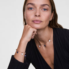 ブラックのVネックトップスを着たモデルが、Blue Spot Heartチャームを含む複数の天然石チャームをあしらったゴールドチェーンのブレスレットとネックレスを身に着けています。ブルーとホワイトのストーンが、胸元と手元に上品なアクセントを添えています。