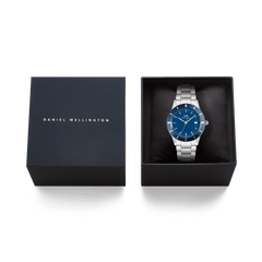 アイコニックなParadigma Link Blue Enamel Silverが、ダークネイビーのブランドロゴ入りギフトボックスに収められています。ブルーエナメルの文字盤とシルバーブレスレットが黒いクッションカバーの上で美しく映え、贈り物にぴったりの上質なパッケージです。