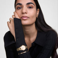 ブラックのニットを着た女性が、Iconic Linkゴールド腕時計とClassic Lumine Braceletを重ね付けした着用イメージです。ゴールドトーンで統一された手元が、ダークな装いに映えます。