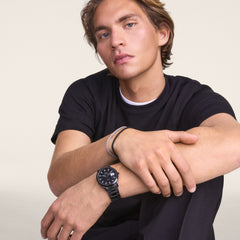 ブラックのTシャツを着た男性が、Daniel WellingtonのIconic Link Automatic Blackを左手首に着用し、ブレスレットと重ね付けしたスタイリングを披露しています。オールブラックのリンクストラップとケースが、カジュアルな装いに自然に馴染んでいます。