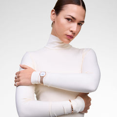 ホワイトのタートルネックを着た女性が、ローズゴールドのメッシュストラップ腕時計とローズゴールドのブレスレット、リングを重ね付けしたスタイリングです。