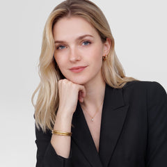 Daniel WellingtonのArch Cuff Goldを2本重ね付けした女性モデルの着用イメージです。黒のテーラードジャケットにゴールドのカフブレスレットとフープピアスを合わせ、洗練されたスタイリングを表現しています。