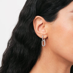 ダニエルウェリントンのCrystal Link Earrings Silverを着用したモデルの耳元アップ。シルバーフープからジルコニアをあしらったオーバルリンクが揺れ、黒髪のウェーブヘアとのコントラストが耳元の輝きを際立たせています。