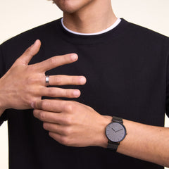 ブラックのクルーネックTシャツに、Daniel WellingtonのClassic Piano Link Onyx Unitone Blackを合わせた男性の手元です。ピアノリンクのオールブラックストラップと文字盤が服装と一体感を演出し、指元にはシルバーのリングをさりげなく添えたスタイリングです。