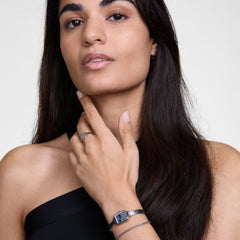 黒のオフショルダートップスを着た女性が、Quadro Mini Reflectionシルバーメッシュ腕時計とClassic Tennis Braceletを重ね付けした手首を顎元に添えた着用イメージです。シルバートーンで統一されたセットが、シンプルな装いに上品なアクセントを添えています。