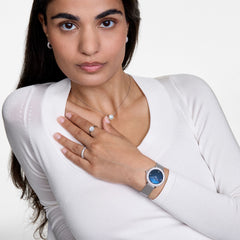 女性がCrystalline Bezel Sterling Blueを左手首に着け、シルバーのネックレスやリング、ピアスとコーディネートした着用イメージです。ブルーマザーオブパールの文字盤が白いニットトップスに美しく映えています。