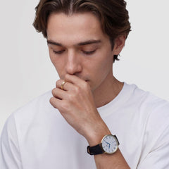 白いTシャツを着た男性が、Daniel Wellingtonのゴールドケースのローマ数字腕時計とゴールドリングを身に着けたスタイリングイメージです。ブラックレザーストラップの腕時計とマットゴールドのリングが、自然に調和しています。