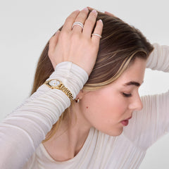 Pavé Ring Gold 3 mmを薬指に着けた女性のスタイリング写真。ゴールドの腕時計やフープピアスと合わせ、クリスタルの煌めきが手元に華やかなアクセントを添えています。